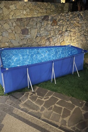 Ein rechteckiger Pool mit blauer Abdeckung, gestützt von weißen Pfählen, vor einer Steinmauer und einer Rasenfläche.