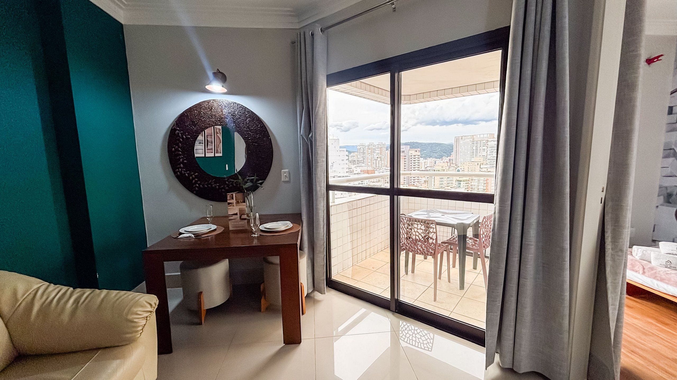 Sala de estar com mesa de jantar, sofá, janela grande com vista para um horizonte urbano e espelho fixado na parede.