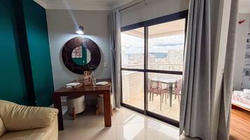 Sala de estar com mesa de jantar, sofá, janela grande com vista para um horizonte urbano e espelho fixado na parede.