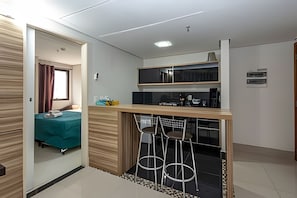 A compact hotel room with a kitchenette, a bed with a teal cover, and a small bar area with stools. - SHB - Beach Vibe Estilo Vista Mar Sacada (Santos)