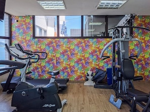 A gym with exercise equipment, a colorful brick wall, and large windows. - SHB - Beach Vibe Estilo Vista Mar Sacada (Santos)