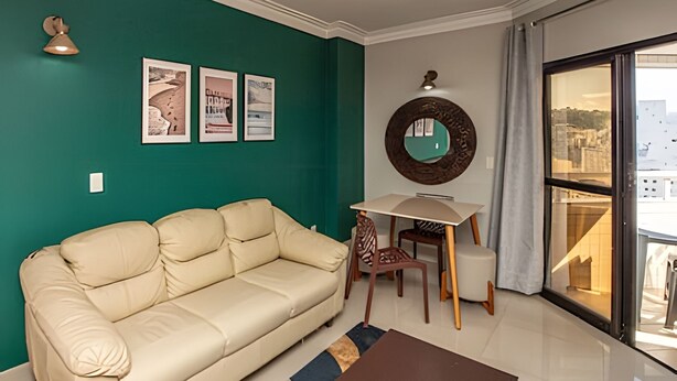 Un salon avec un canapé beige, une petite table et un fauteuil. Trois tableaux encadrés ornent le mur, ainsi qu’un miroir rond et une fenêtre donnant sur des immeubles.