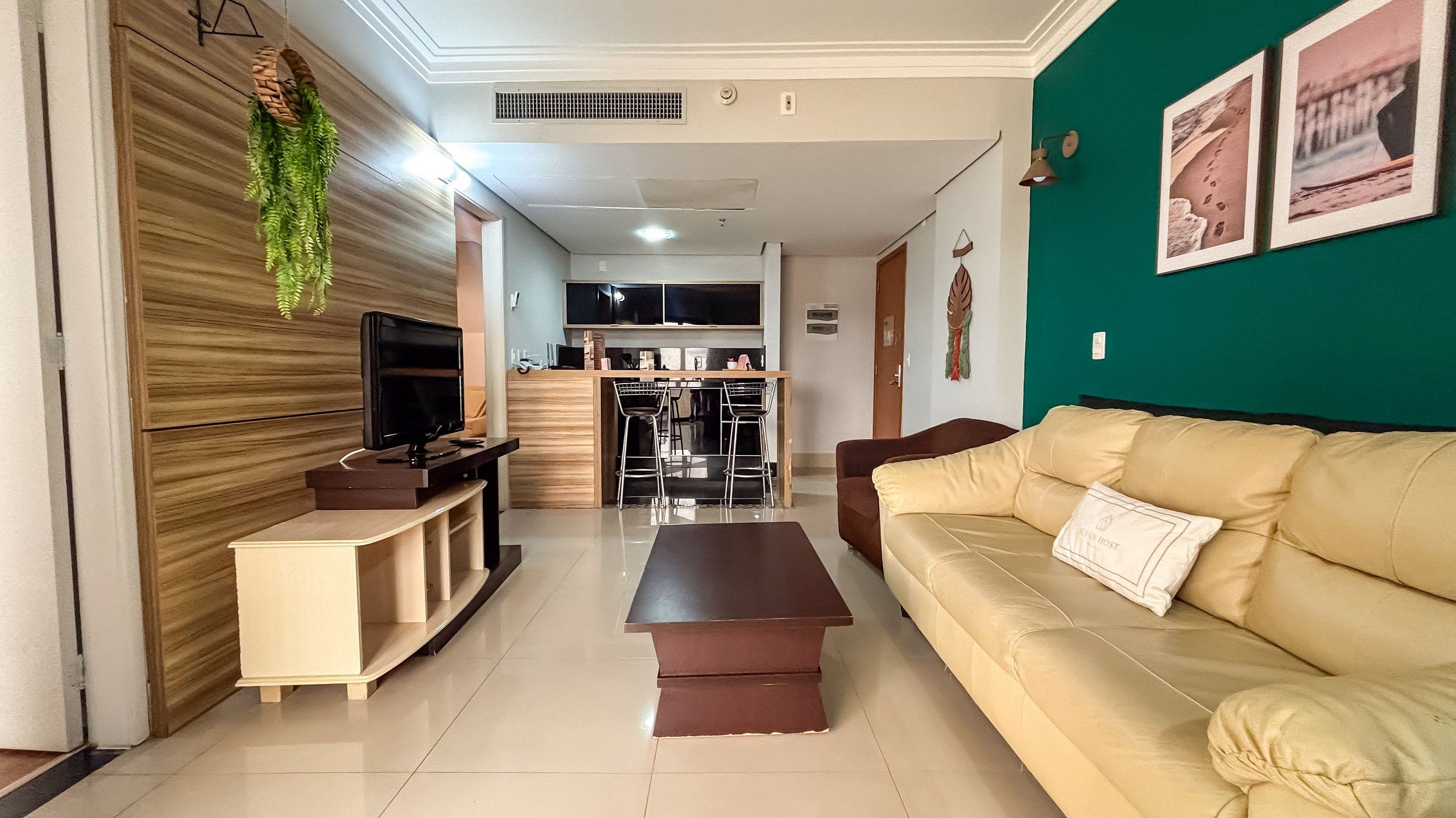Una sala de estar con un sofá color beige, un televisor sobre un mueble bajo y una cocina al fondo.