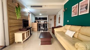 Una sala de estar con un sofá color beige, un televisor sobre un mueble bajo y una cocina al fondo.