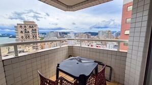 A balcony with a table and chairs, overlooking a cityscape with buildings and a body of water. - SHB - Beach Vibe Estilo Vista Mar Sacada (Santos)