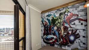Une chambre avec une fresque murale représentant des super-héros, un lit et une fenêtre donnant sur un paysage urbain.