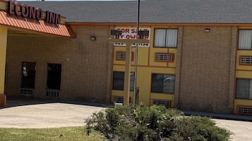 Un motel de varios pisos con fachada amarilla y marrón, un área de estacionamiento techada y un letrero que indica disponibilidad para la venta.