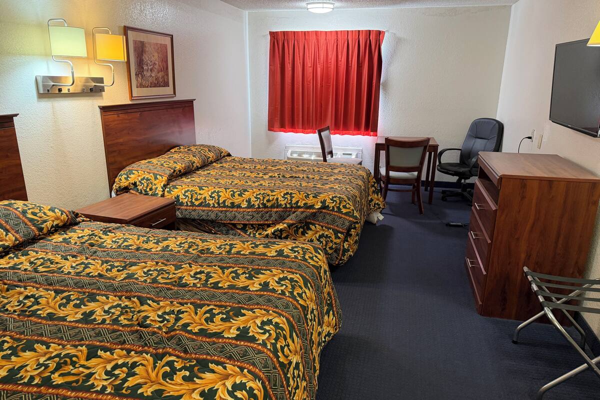 A hotel room with three beds, a desk, a chair, a TV, and a window with red curtains.