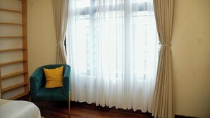 A room with a bed, a chair with a yellow pillow, a wooden shelf, and a large window with sheer curtains. - Sophies Garden Hotel (Makutano)