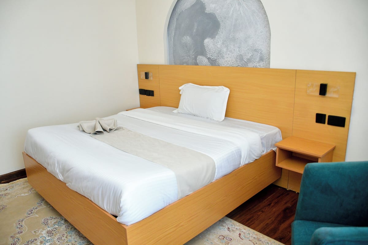 A neatly made bed with a wooden headboard, a bedside table, and a chair in a room with a patterned rug.