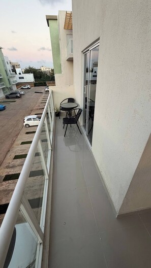 A balcony with a table and chairs, a view of a parking lot, and a building with a green facade. - Welcome to Your Home 15 Minutes From the Airport (Santo Domingo Este)