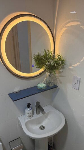 A bathroom with a round mirror, a sink, a vase with a plant, and a soap dispenser. - Welcome to Your Home 15 Minutes From the Airport (Santo Domingo Este)