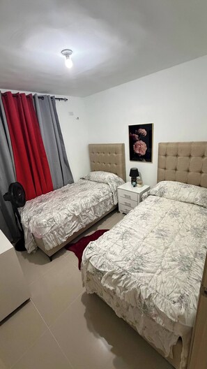 A bedroom with two beds, a nightstand, a lamp, a red curtain, and a ceiling light. - Welcome to Your Home 15 Minutes From the Airport (Santo Domingo Este)