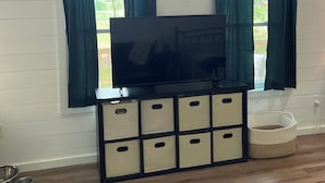 A room with a TV on a black stand, two windows with curtains, a white wall, and a wooden floor.