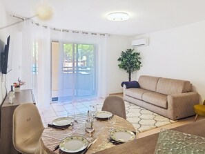 Et spiseområde med et bord dækket til fire personer, en sofa, en potteplante og en skydedør til terrassen.