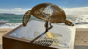 En metalkunstinstallation af en fugl på en reflekterende overflade med kystlandskab i baggrunden.