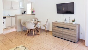 Et moderne køkken med hvide skabe, et spiseområde med et rundt bord og stole, et TV ophængt på væggen og en trækommode.