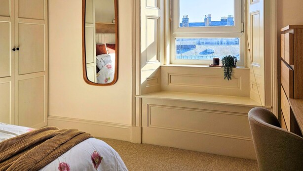 Hotelkamer met een groot raam, een bed met bloemenbedekking, een stoel en een spiegel.