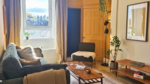 Een woonkamer met een bank, fauteuil, salontafel en uitzicht op gebouwen door een raam.