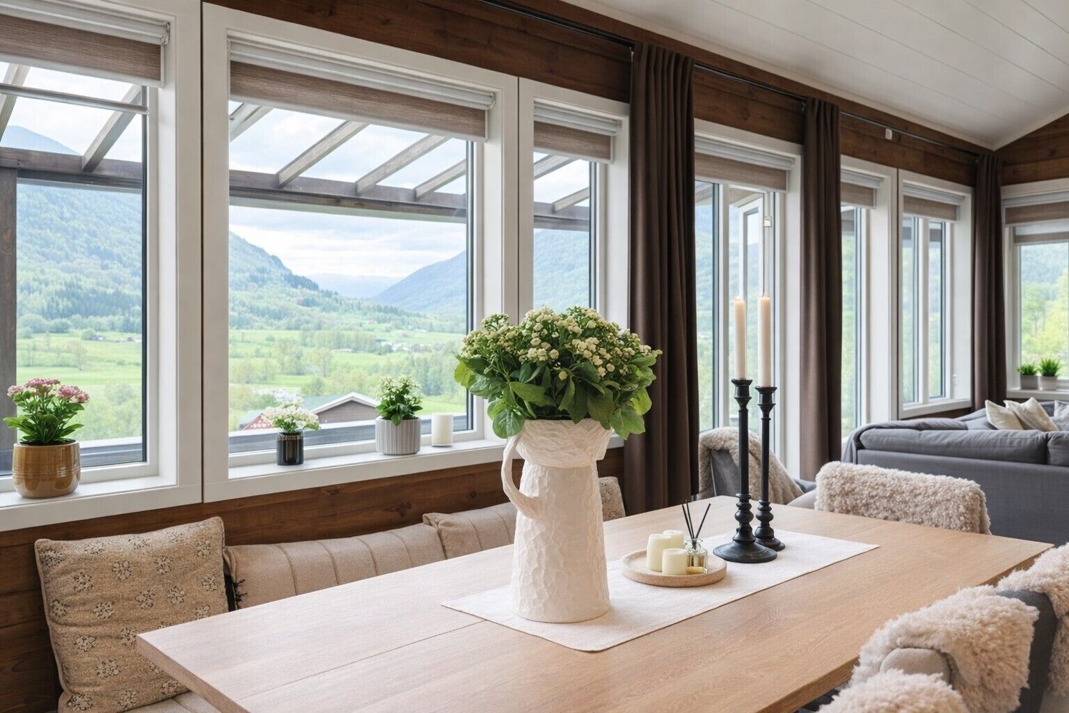 A room with a wooden table, a vase with flowers, a candle holder, and a view of mountains through large windows.