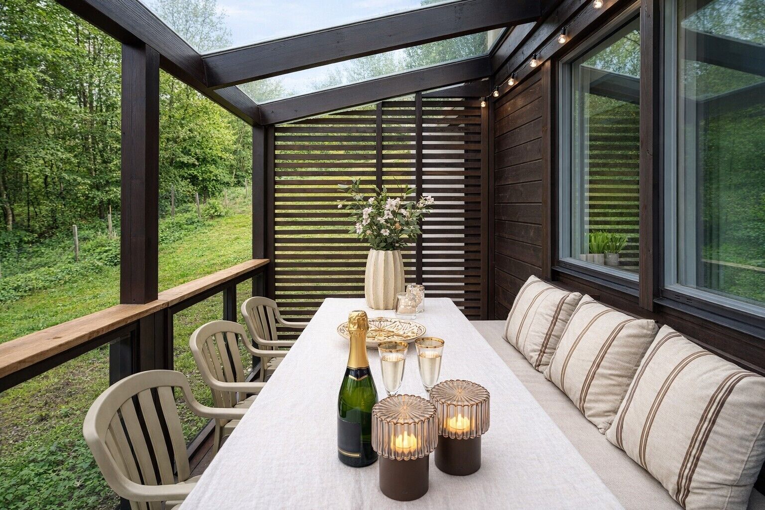 A covered outdoor seating area with a table, chairs, a bottle of champagne, candles, and a vase of flowers.