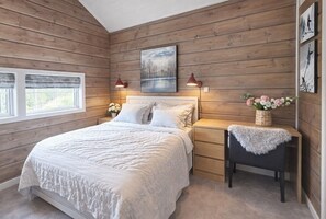 Quarto com parede de madeira, cama, mesas de cabeceira, criadeira, abajur e um vaso com flores.