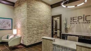 Ein Empfangsbereich mit Steinwand, einer Rezeption, einem Stuhl und einem an der Wand befestigten Schild mit der Aufschrift 'THE EPIC SPA & FITNESS'.