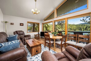 A spacious living room with a wooden coffee table, leather sofas, and a dining area with chairs. Large windows offer a view of a lake and forest. - Waterfront Sandbar Point w/ Dock Access & Nearby Activities (Hayden)