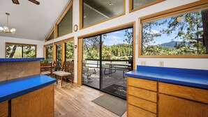 Cozinha com armários de madeira e bancada azul, área de refeições com mesa e cadeiras, e grandes janelas com vista para árvores e montanhas.