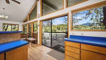 Une cuisine avec des meubles en bois et un plan de travail bleu, un coin repas avec une table et des chaises, et de grandes fenêtres offrant une vue sur les arbres et les montagnes.