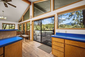 A kitchen with wooden cabinets and a blue countertop, a dining area with a table and chairs, and large windows offering a view of trees and mountains. - Waterfront Sandbar Point w/ Dock Access & Nearby Activities (Hayden)
