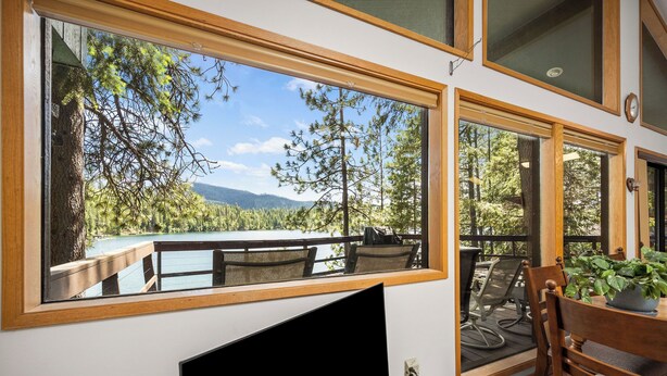 Quarto com uma grande janela com vista para um lago e uma floresta, uma mesa de jantar com cadeiras e uma televisão de tela plana fixada na parede.