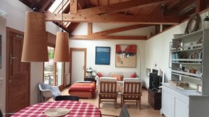 Une pièce avec un plafond en bois, une table à manger recouverte d’une nappe à carreaux rouges et blancs, deux chaises en bois, un canapé et une bibliothèque.