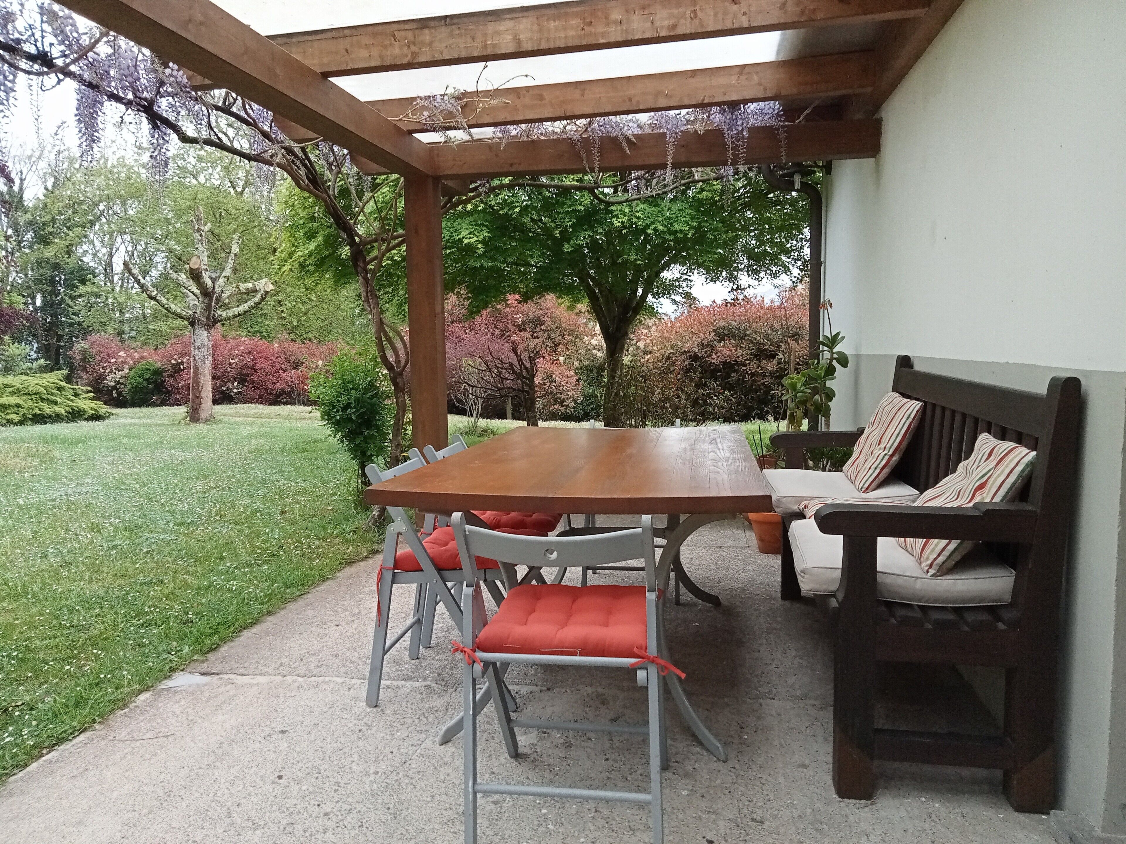 Een overdekt terras met een houten tafel en stoelen, een bankje en uitzicht op een tuin met bomen en struiken.