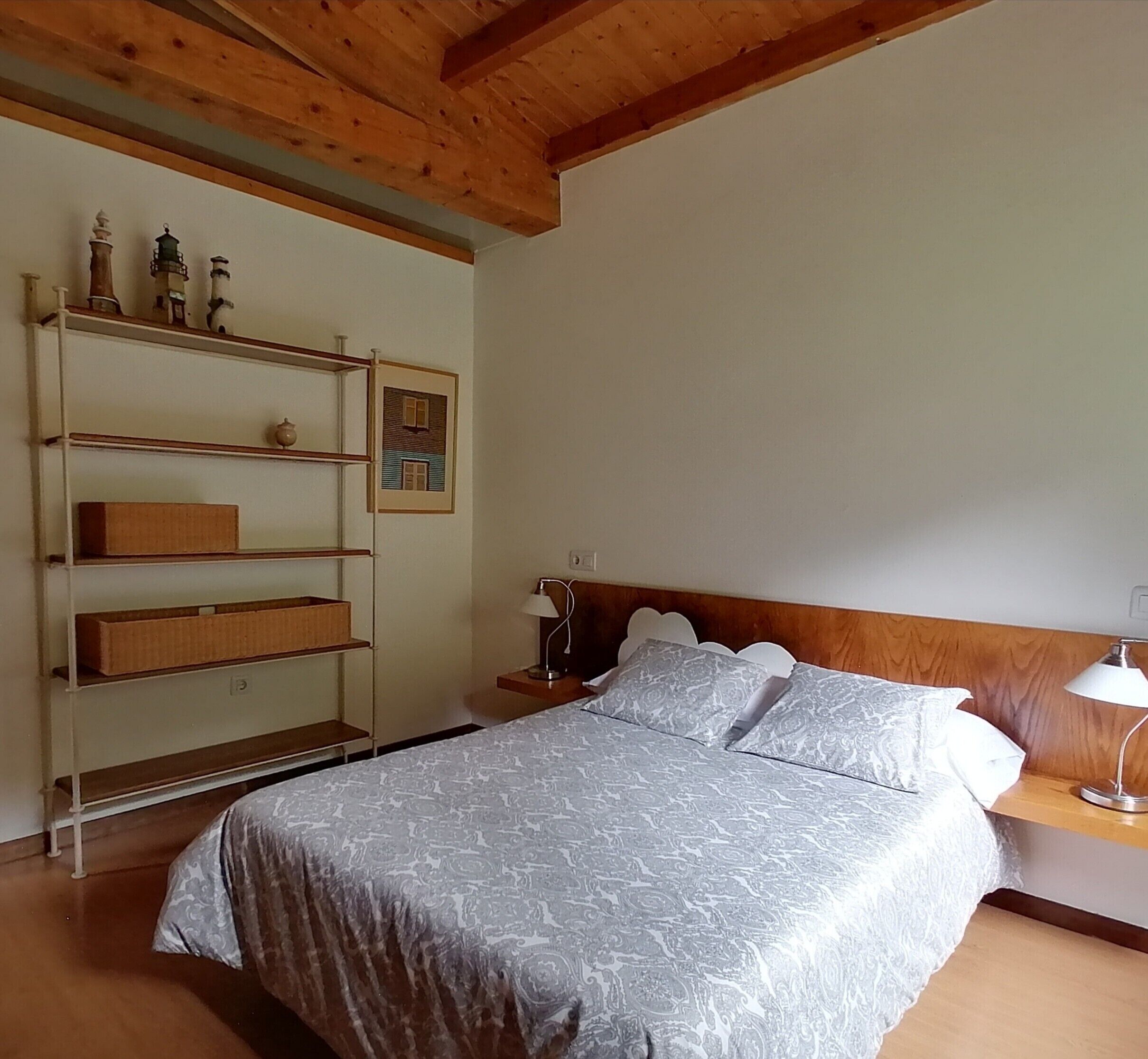 Een slaapkamer met een bed, nachtkastje, lamp, plank met decoratieve objecten en een ingelijste foto aan de muur.