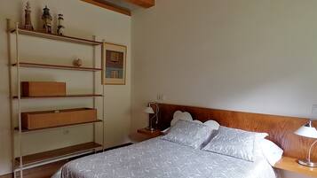 Een slaapkamer met een bed, nachtkastje, lamp, plank met decoratieve objecten en een ingelijste foto aan de muur.