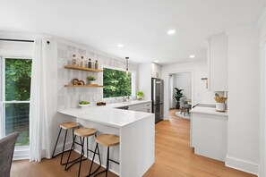 A modern kitchen with a large island, bar stools, built-in shelves, and a view of greenery outside.