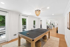 A ping pong table in a bright room with large windows, a chandelier, and a kitchen in the background.