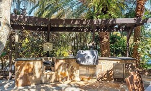 Un espace barbecue extérieur doté de plans de travail en pierre et surmonté d’une pergola.