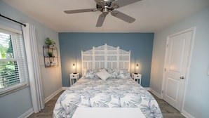 A bedroom with a bed, a ceiling fan, bedside lamps, a nightstand, a bench, a decorative headboard, and a door. - Relax at our fully renovated peaceful beach condo in Satellite Beach (Satellite Beach)