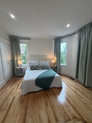 Ein Schlafzimmer mit einem großen Bett, zwei Nachttischen mit Lampen, einem Fenster mit Vorhängen und einem Holzboden.