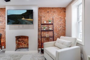 Ein Wohnzimmer mit Kamin aus Ziegelsteinen, einem an der Wand befestigten Fernseher, einem weißen Sofa und einem Holzregal mit Dekoration.