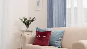 Un salon avec un canapé beige, un coussin rouge orné d’un logo, un coussin bleu, une petite table avec une plante et un tableau encadré accroché au mur.
