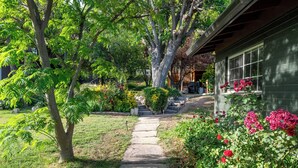 Um caminho de pedras leva a uma casa com fachada verde, cercada por vegetação exuberante e plantas floridas.