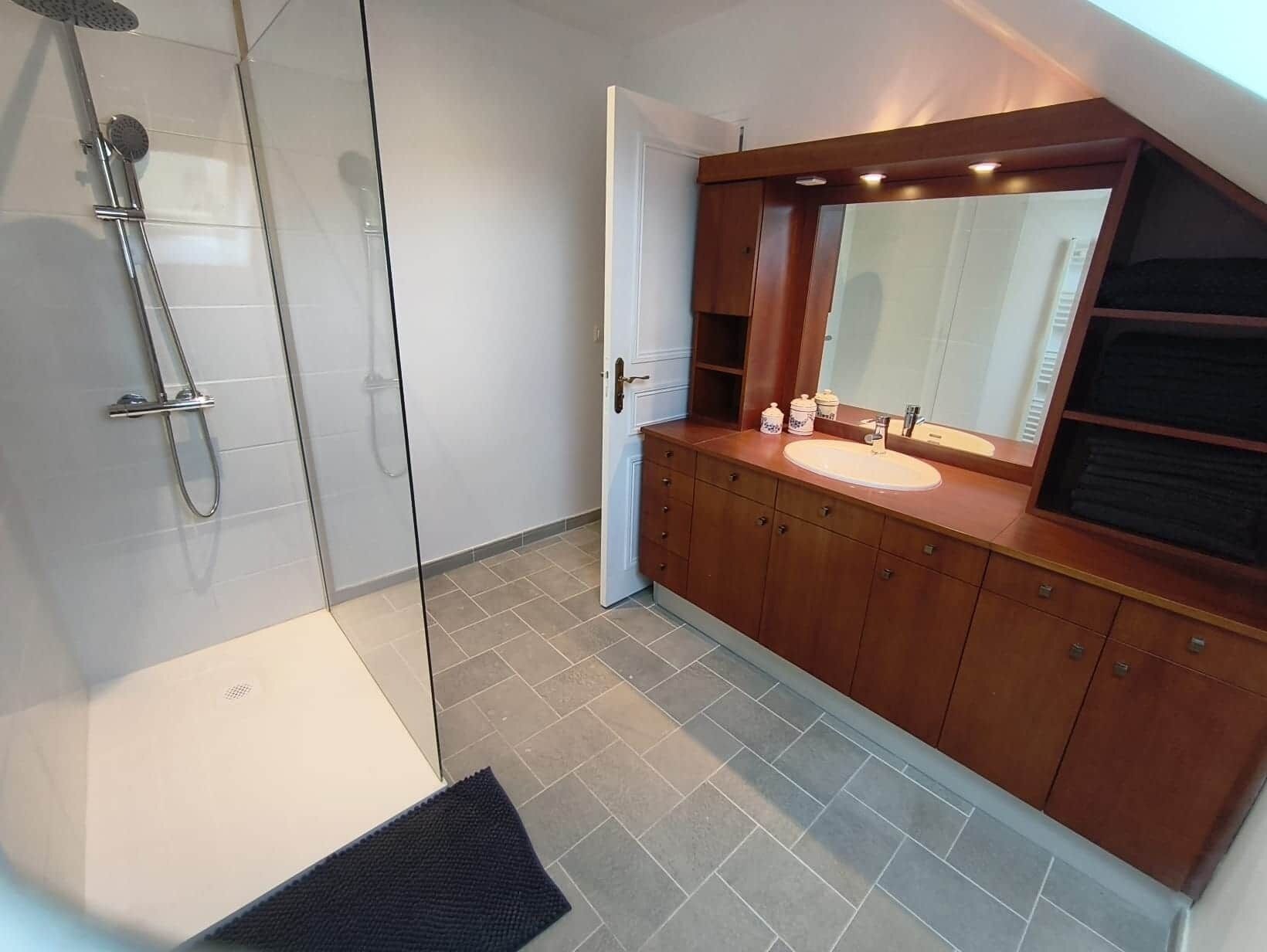 Een moderne badkamer met een glazen douchecabine, een houten wastafelmeubel met een wastafel en een grote spiegel erboven.
