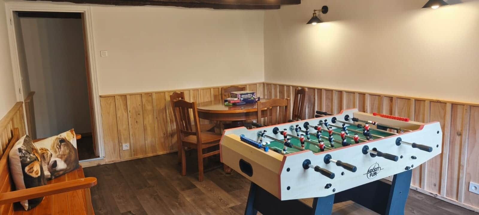 Una habitación con mesa de futbolín, un banco de madera, una silla y una pequeña mesa con una caja encima.