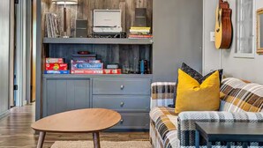 A living room with a wooden wall, a sofa with a plaid cover, a coffee table, a lamp, a guitar, and a bookshelf with various items. - Creekside retreat + hot tub near Sedona (Cornville)