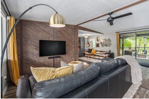 A modern living room with a leather sofa, a flat-screen TV, a ceiling fan, and a large window. - Creekside retreat + hot tub near Sedona (Cornville)
