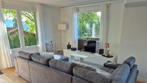 A modern living room with a grey sectional sofa, a flat-screen TV, a white console table, and a large window with a view of greenery. - Apartment Sunshine lake and mountain view with 60 sqm terrace facing south and east (Millstatt)