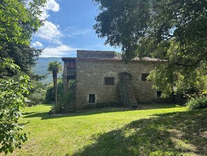 A stone building with a tiled roof, surrounded by greenery and a clear sky.